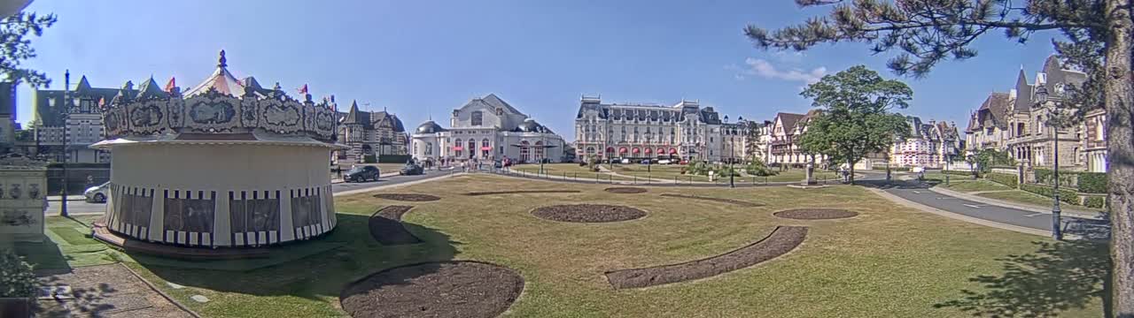 A carousel sits in a grassy town square in front of several large buildings on a sunny day.