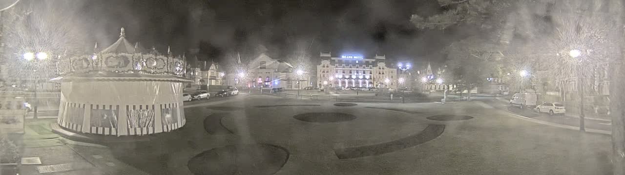 A snow-covered town square at night features a carousel, several buildings, and parked cars under dim streetlights.