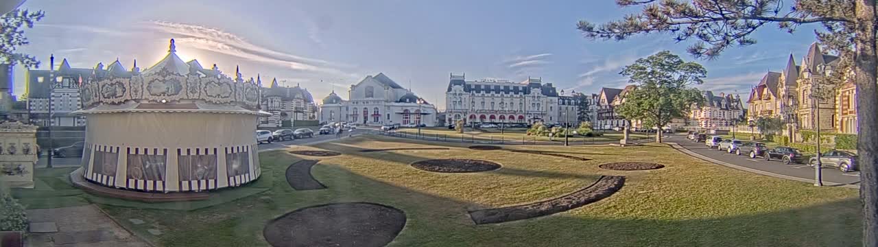 A carousel sits near a grassy park in front of several large buildings under a mostly sunny sky.