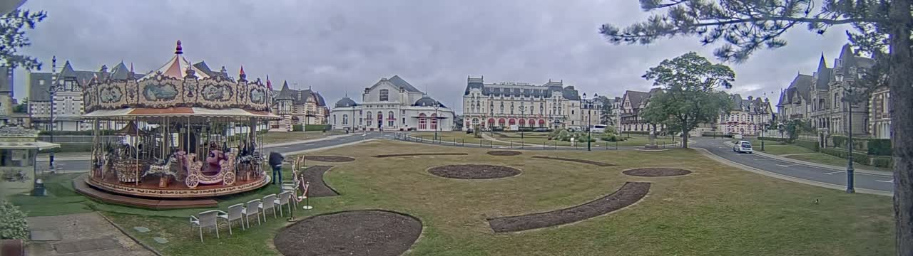 A carousel sits in a grassy park with landscaped gardens in front of several large buildings under a cloudy sky.