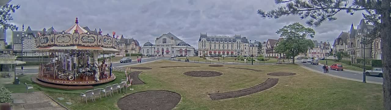 A carousel sits in a grassy town square, overlooked by several large buildings under an overcast sky.