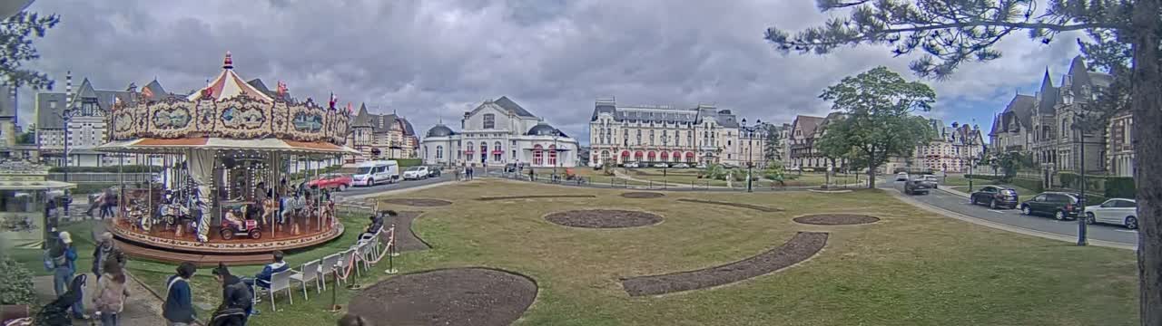 A carousel sits in a town square with several large buildings in the background under a cloudy sky.