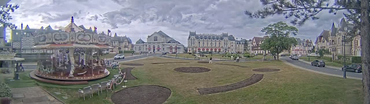 A carousel sits in a grassy town square under a cloudy sky, with several large buildings and a few cars visible in the background.