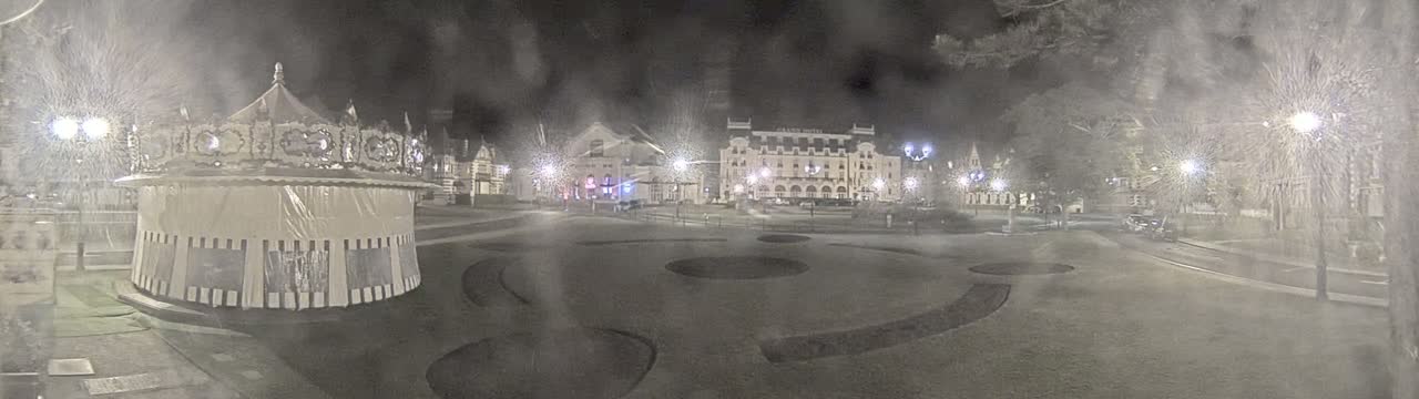 A snow-covered carousel sits in a town square at night, near several buildings, under streetlights.