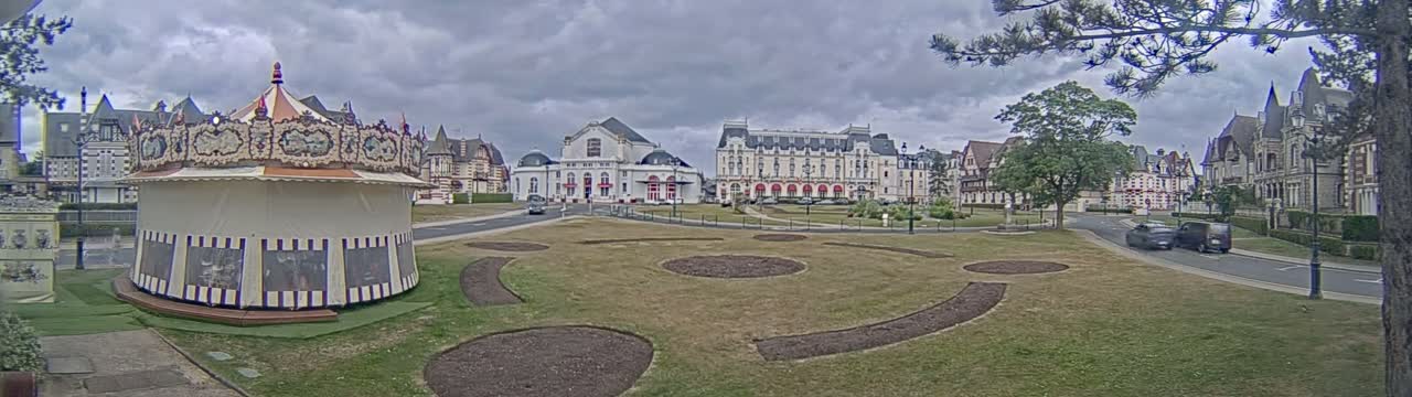 A carousel sits in a grassy town square, overlooked by several large buildings under a cloudy sky.