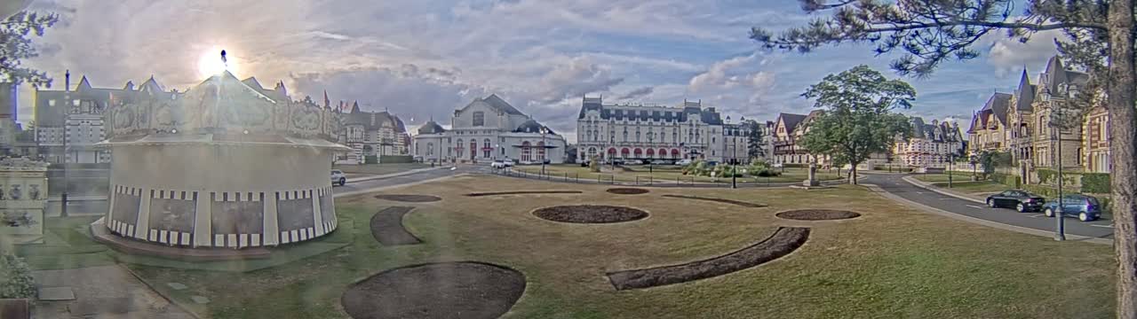 A carousel sits in a grassy park with patterned flower beds in front of several large buildings under a partly sunny sky.