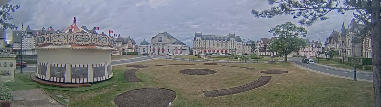 A carousel sits in a grassy town square under an overcast sky, with several large buildings and trees surrounding it.