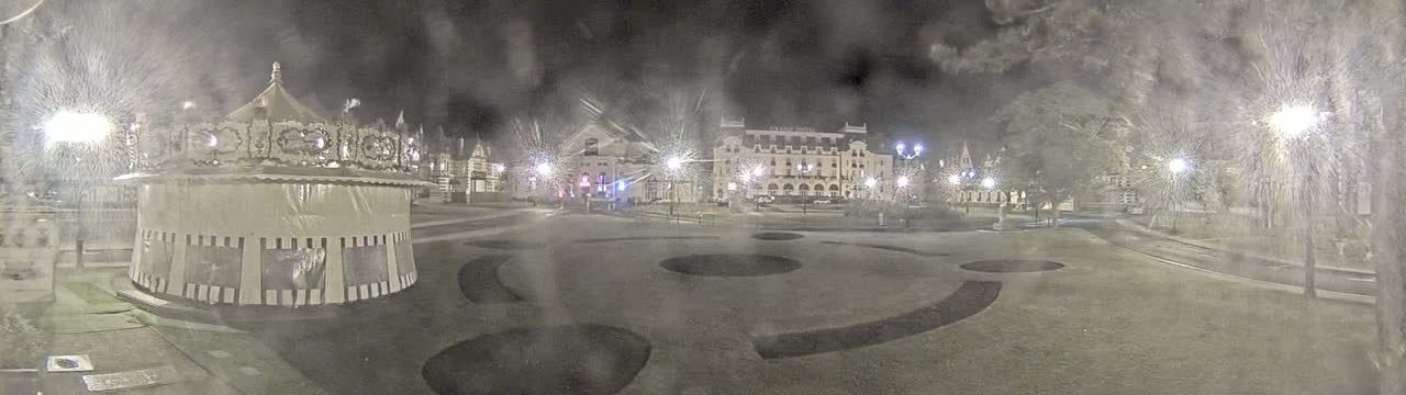 A carousel sits in a snow-covered town square at night, with several buildings and trees visible in the background under streetlights.