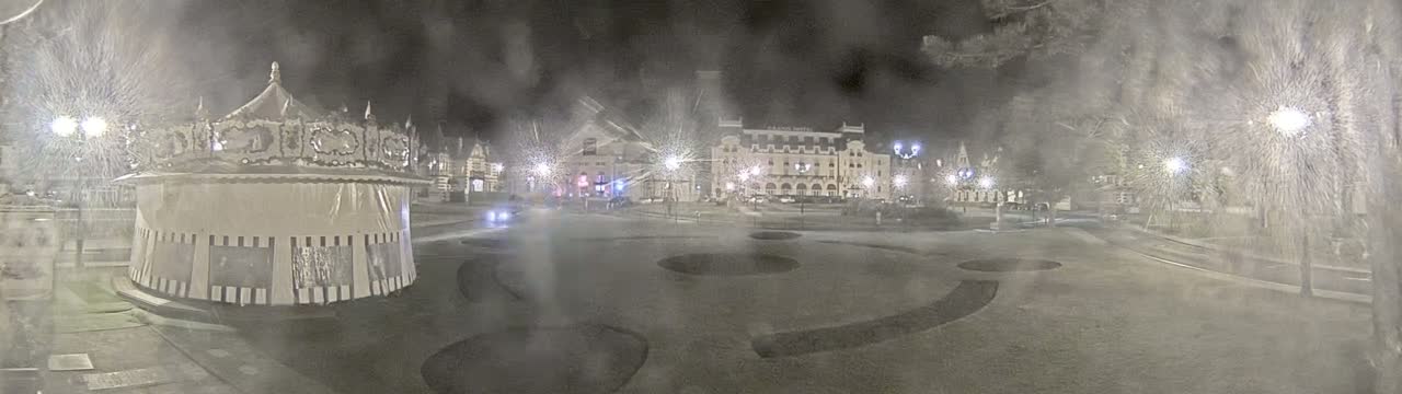 A snow-covered carousel sits in a town square at night, with a large building visible in the background under heavy snowfall.