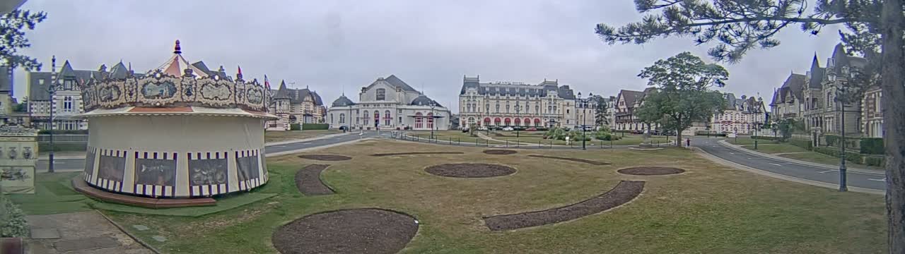 A carousel sits in a grassy town square in front of several large buildings under an overcast sky.