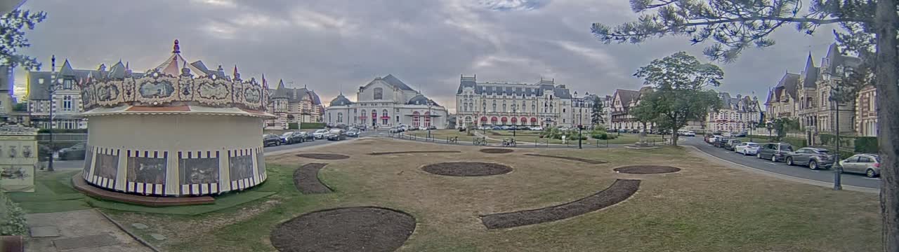 A carousel sits in a town square, surrounded by several large buildings and a sparsely vegetated lawn under a cloudy sky.