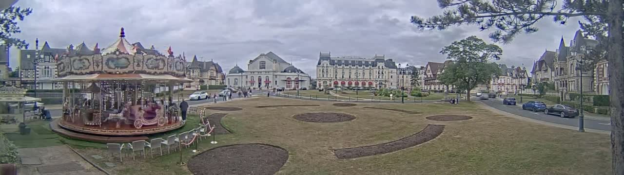 A carousel sits in a town square with several buildings, including a large hotel, under an overcast sky.