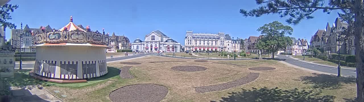 A carousel sits in a grassy town square on a sunny day, with several large buildings, including a hotel, in the background.