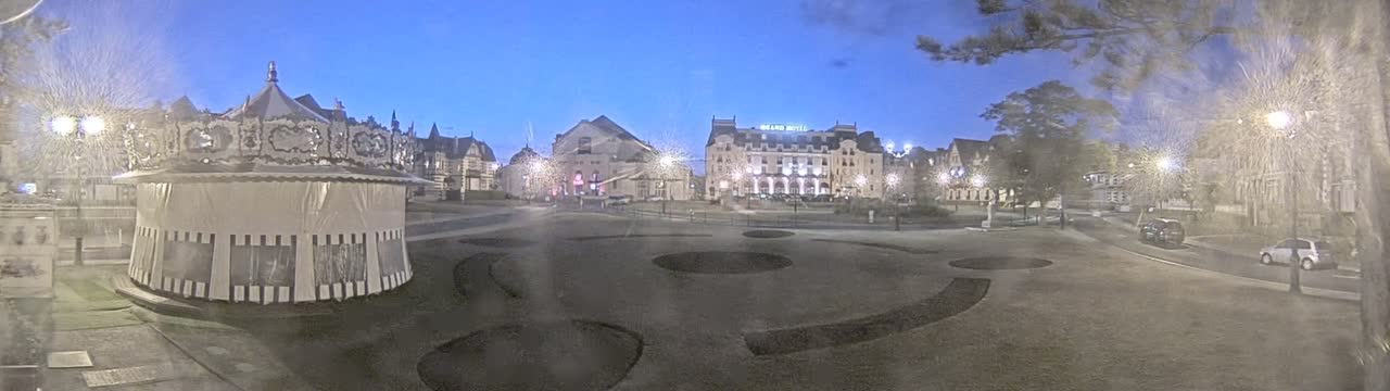A carousel sits in a town square at twilight, surrounded by buildings, trees, and a few parked cars.