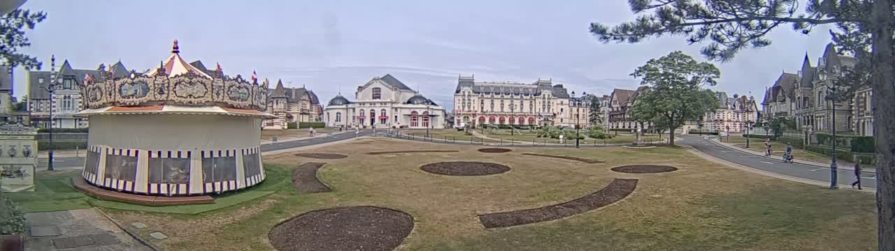 A carousel sits in a grassy town square, in front of several large buildings under a mostly clear sky.