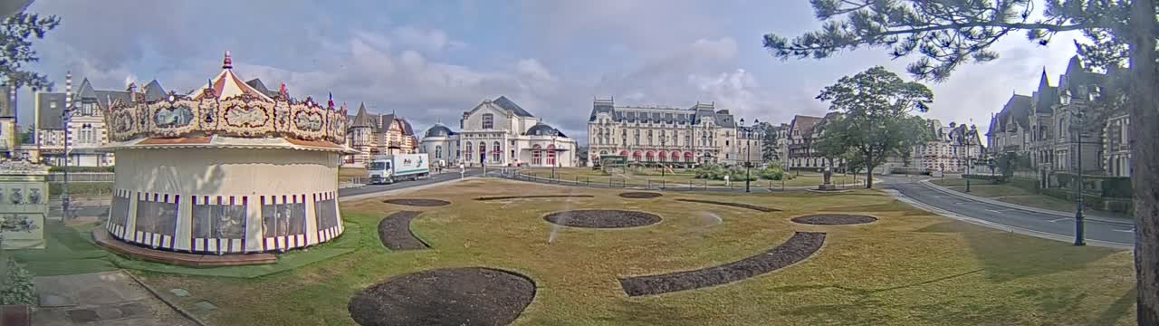 A carousel sits in a grassy park area in front of several large buildings under a partly cloudy sky.