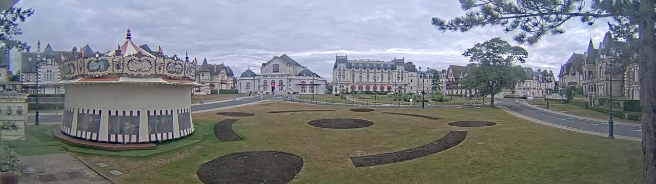 A carousel sits in a grassy park in front of several large buildings under an overcast sky.
