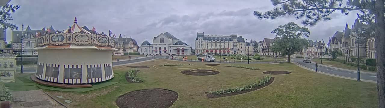 A carousel sits in a grassy town square under an overcast sky, in front of several large buildings.