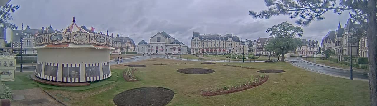 A carousel sits in a grassy square in front of several large buildings on an overcast day.