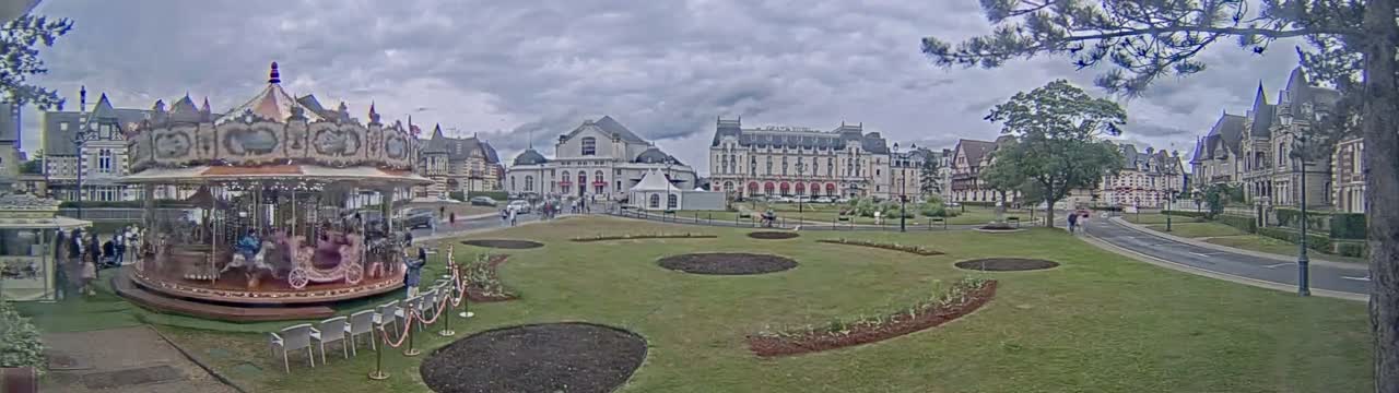 A carousel sits in a grassy town square, surrounded by ornate buildings under a cloudy sky.