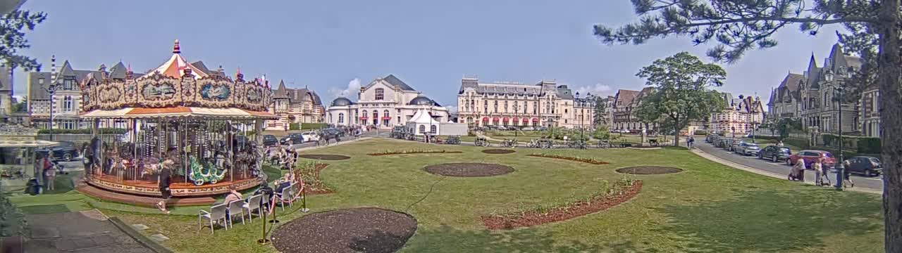 A carousel sits in a grassy town square on a sunny day, surrounded by several buildings and people, with cars parked along a nearby street.