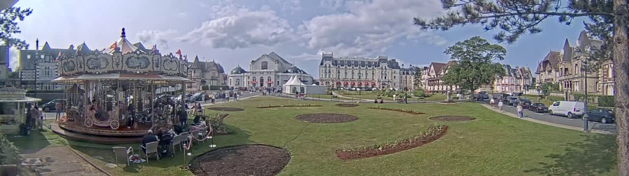 A carousel sits in a grassy town square, surrounded by several buildings under a partly cloudy sky.