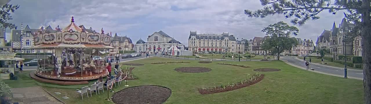 A carousel sits in a grassy town square under cloudy skies, near several large buildings and a road.