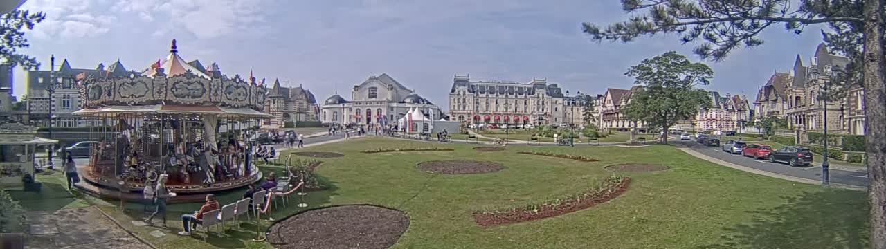A carousel sits in a grassy town square bordered by several large buildings under a partly cloudy sky.