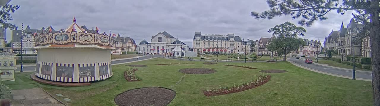 A carousel sits in a grassy park area in front of several large buildings under an overcast sky.