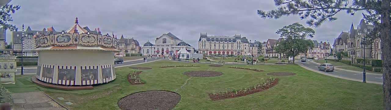 A carousel sits in a grassy park area next to several large buildings under an overcast sky.