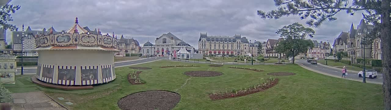 A carousel sits in a grassy town square, surrounded by several large buildings under an overcast sky.