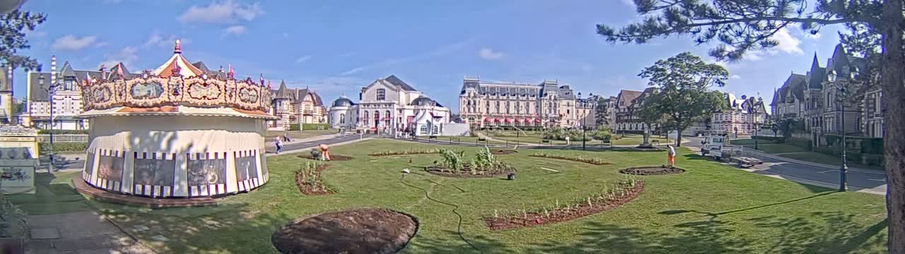 A carousel sits in a grassy town square on a sunny day in front of several large buildings.