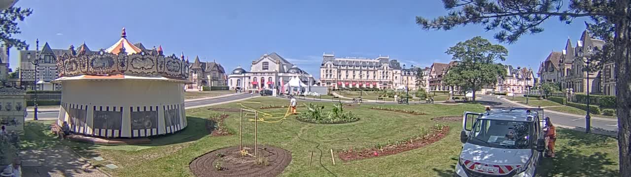 A carousel sits in a grassy town square on a sunny day, with several buildings, including a large hotel, visible in the background.