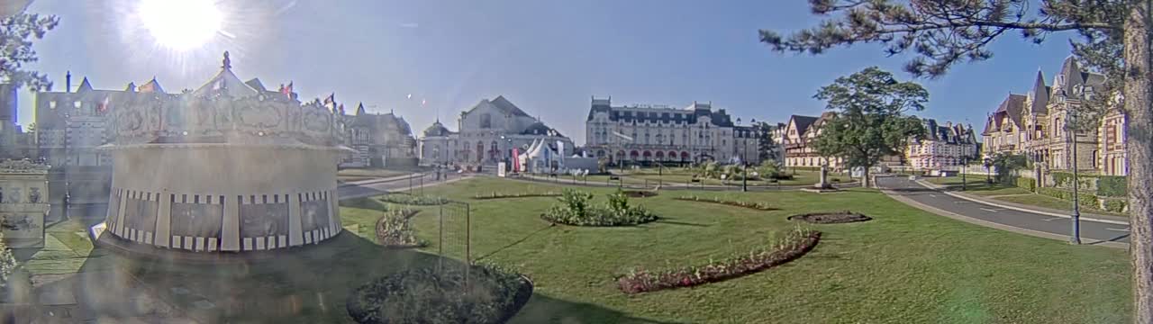 A sunny day reveals a picturesque town square with a carousel, manicured lawns, and several grand, ornate buildings in a European architectural style.