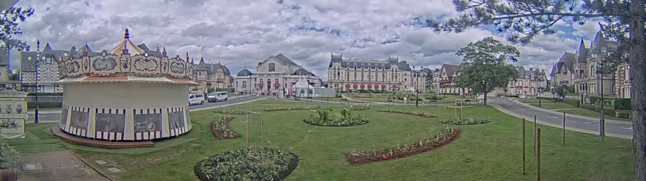 A carousel sits in a grassy park in front of several large buildings under a partly cloudy sky.