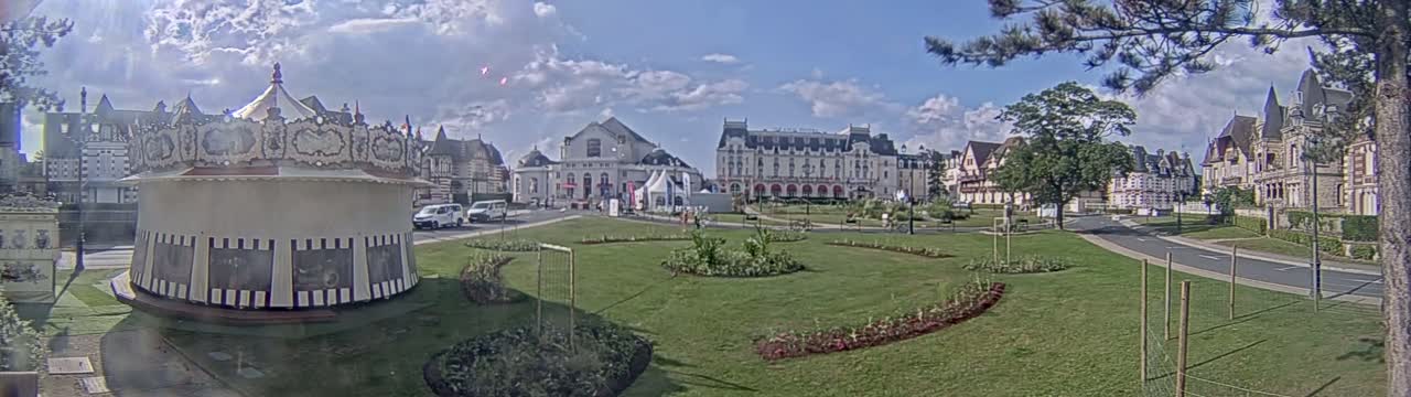 A carousel sits in a grassy park next to several large buildings on a partly cloudy day.