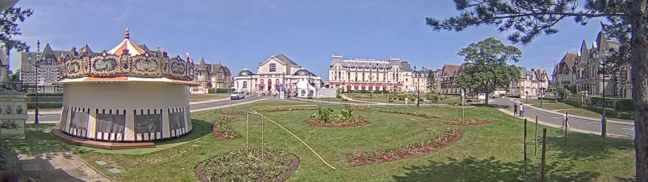 A carousel sits in a grassy town square next to several large buildings under a sunny sky.