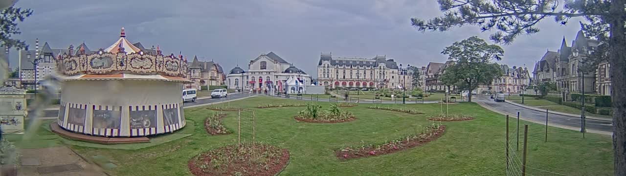 A carousel sits in a grassy park next to a street lined with large buildings under an overcast sky.