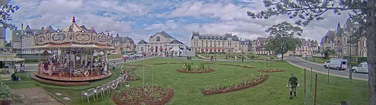 A carousel sits in a grassy park area in front of several large buildings under a partly cloudy sky.
