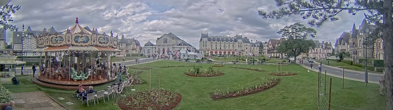 A carousel sits in a grassy park surrounded by several large buildings under a cloudy sky.