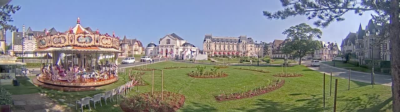 A carousel sits in a grassy town square on a sunny day, surrounded by several buildings of varying architectural styles.