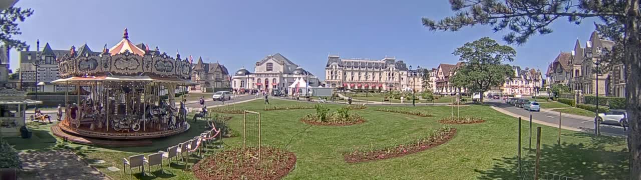 A carousel sits in a grassy town square on a sunny day, surrounded by several buildings and a few parked cars.