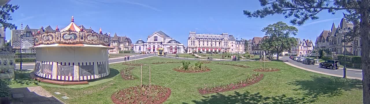 A carousel sits in a grassy park in front of several large buildings on a sunny day.
