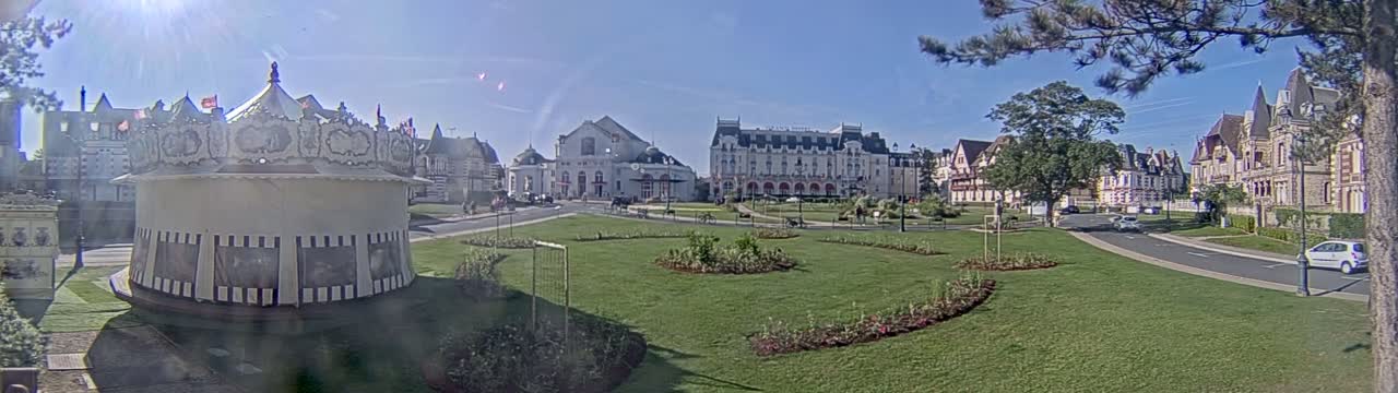 A carousel sits in a grassy park area in front of several large buildings on a sunny day.