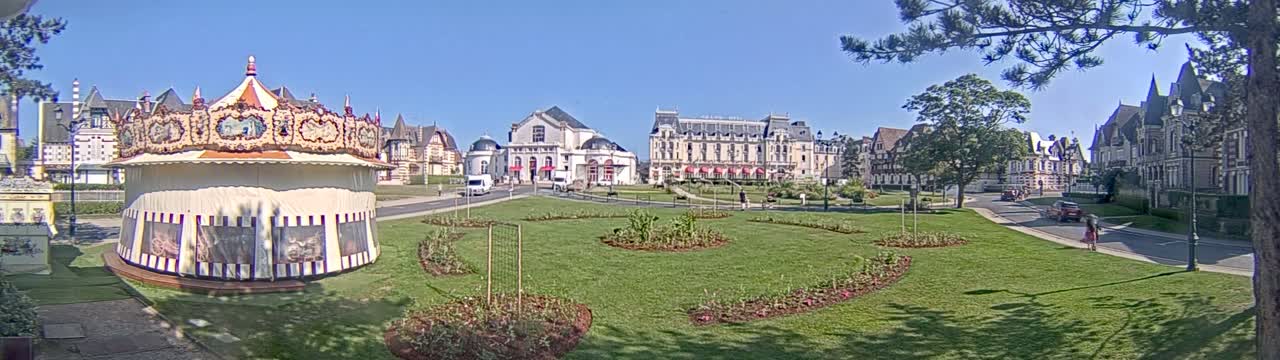 A carousel sits in a grassy park in front of several ornate buildings on a sunny day.