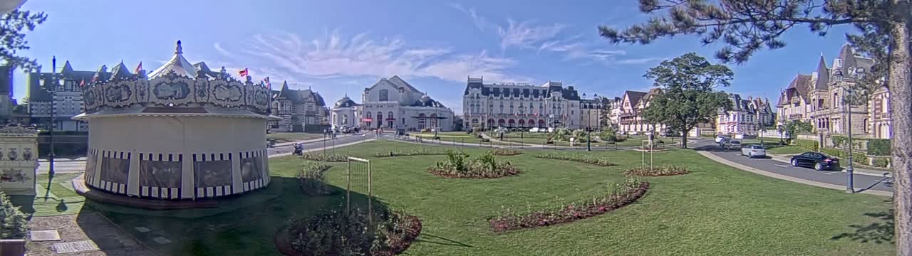 A carousel sits in a grassy town square, in front of several large buildings, under a mostly sunny sky.