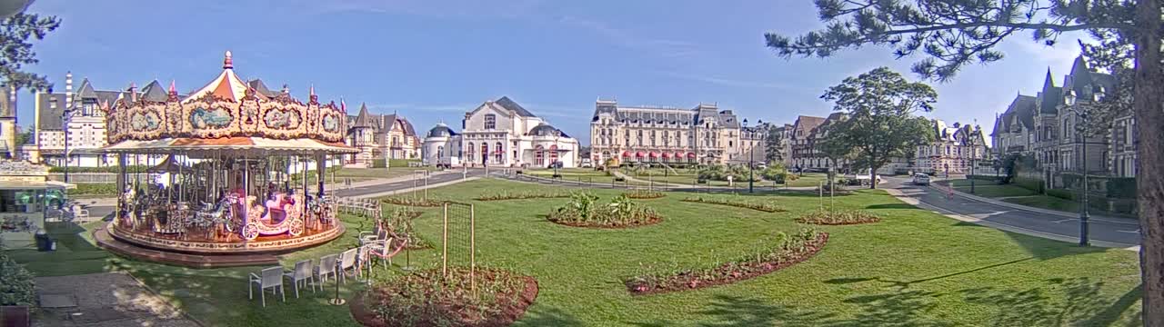 A carousel sits in a grassy town square, surrounded by several ornate buildings under a sunny sky.