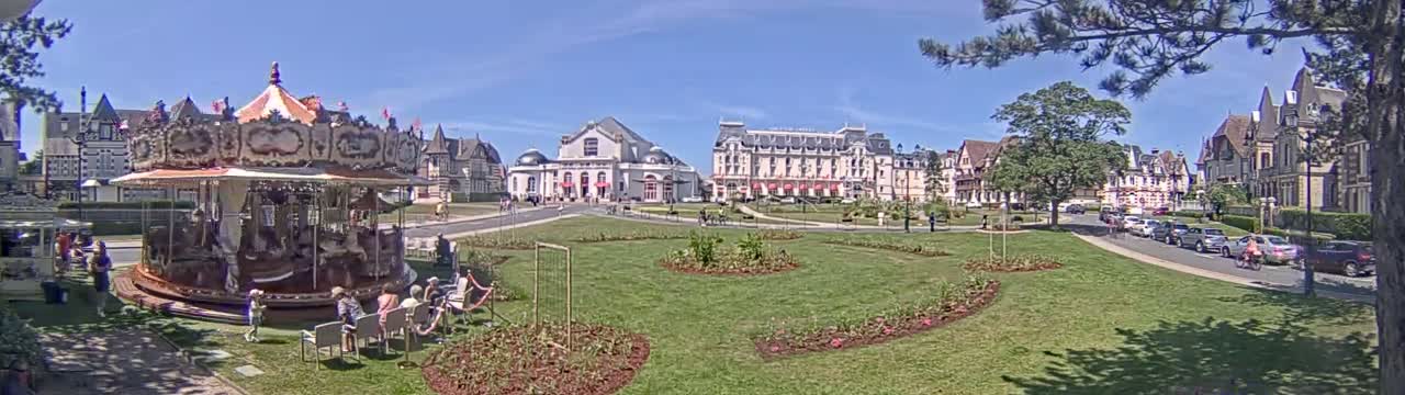 A carousel sits in a grassy town square on a sunny day, with several buildings, including a large hotel, and parked cars lining a nearby street.