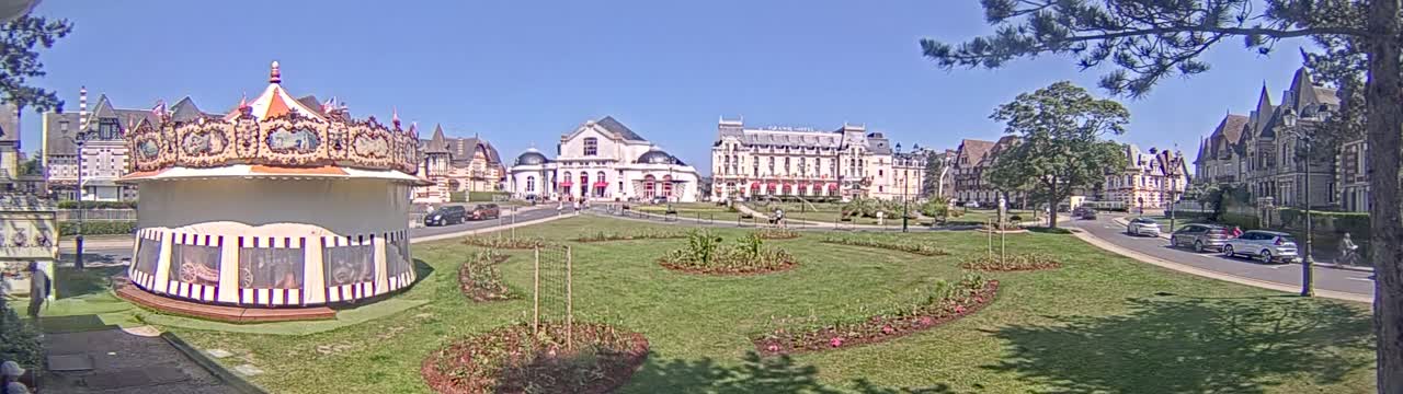 A carousel sits near a grassy park in front of several large buildings on a sunny day.