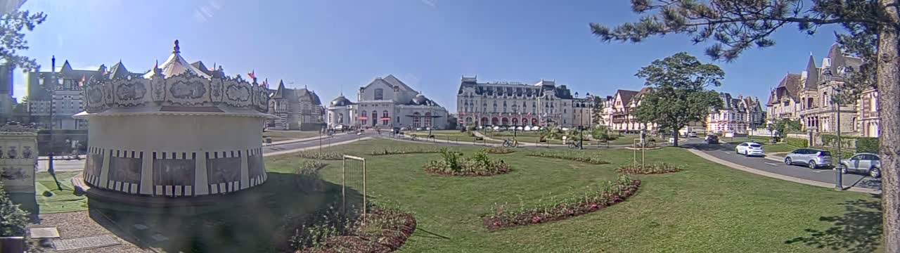 A carousel sits in a grassy park area in front of several large buildings, on a sunny day.